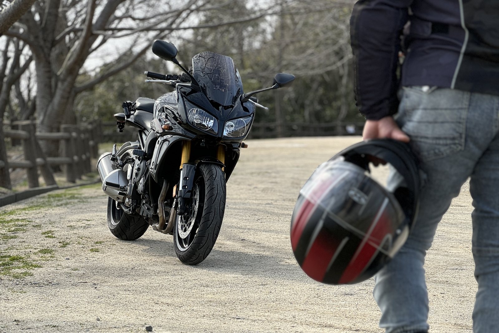 並木道に停めた黒い大型バイクと、赤黒のヘルメットを提げて歩くロイ